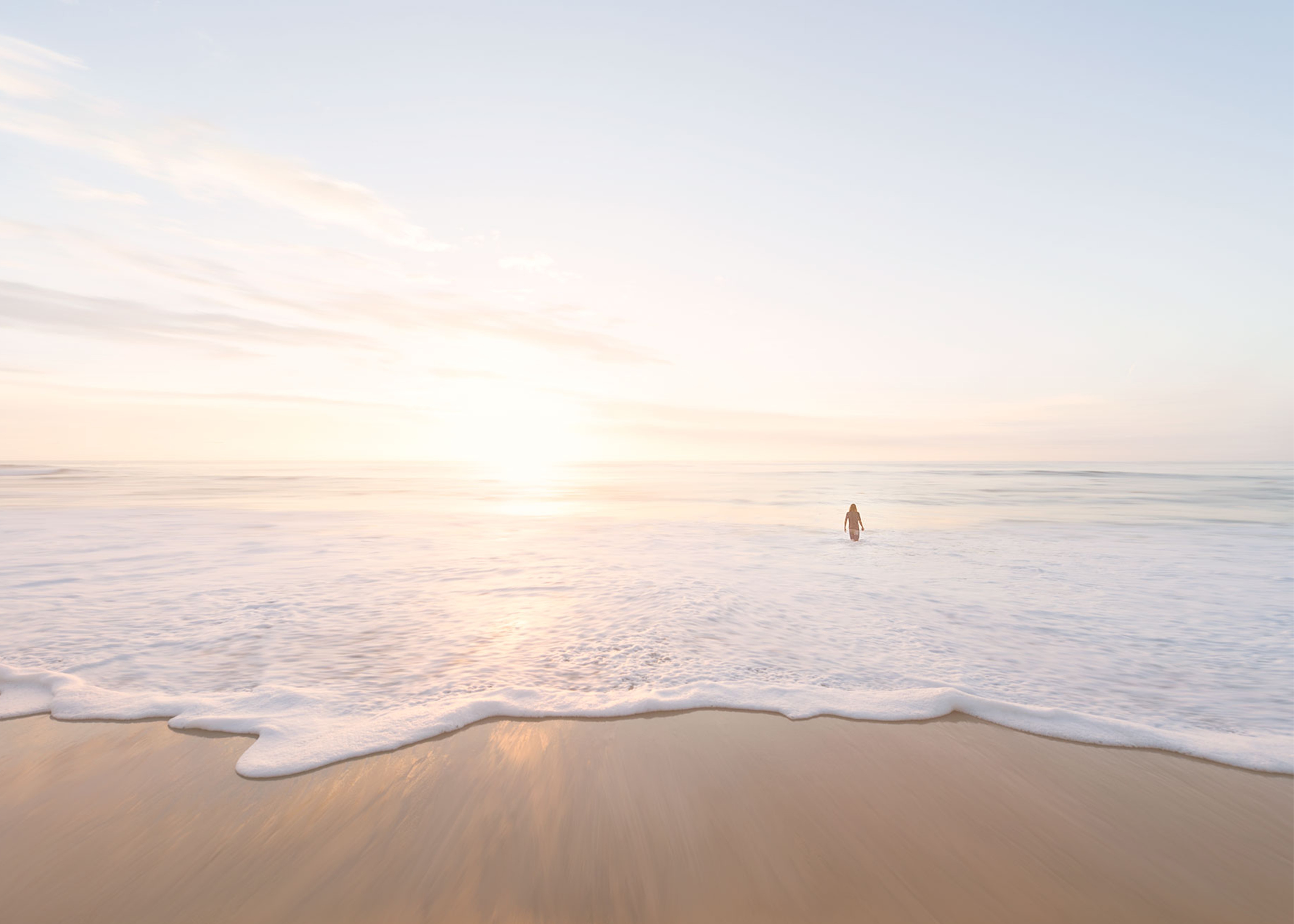 A person standing in shallow ocean water facing a bright sunrise on a sandy beach